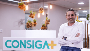 Victor Loyola explica empréstimo consignado. Ele está sentado ao lado lo logo da Consiga+. Ele está de camisa branca e de braço cruzado