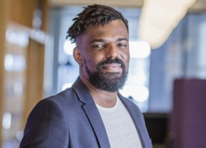 Uribe Teófilo está de cabelo trançado, com barba, vestindo um paletó azul sobre uma camiseta branca