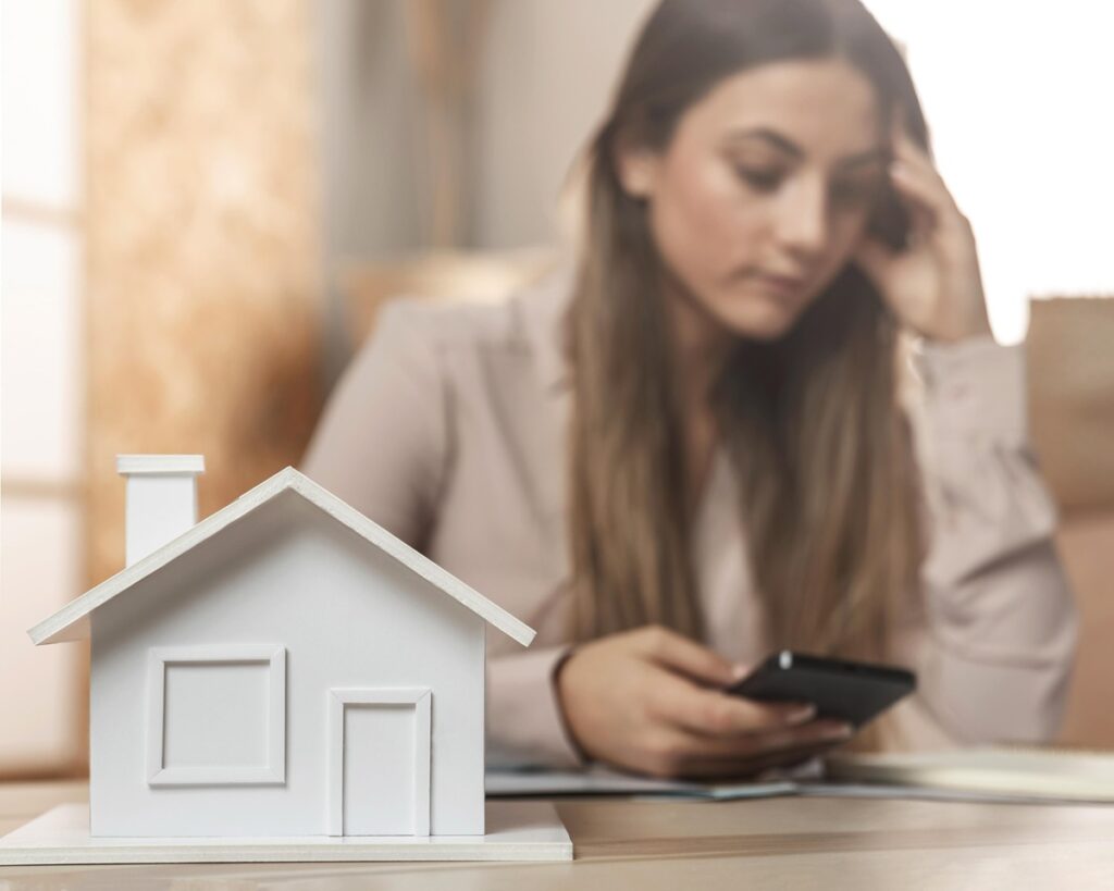 Moça de cabelos loiros e longos olha celular preocupada e à sua frente há uma miniatura de uma casa