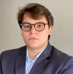 O advogado Luan Mazzalli Braughat tem cabelos castanhos claros, e jove3m, usa óculos e está de terno