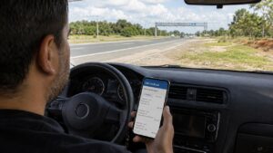 motorista sentado no carro olhando o aplicativo da CNH no celular e logo a frente um pórtico de pedágio free flow